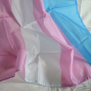 May include: A transgender pride flag made of fabric with light blue, pink, and white stripes. The flag is draped over a white surface.
