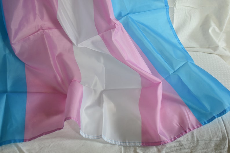 May include: A transgender pride flag made of fabric with light blue, pink, and white stripes. The flag is draped over a white surface.
