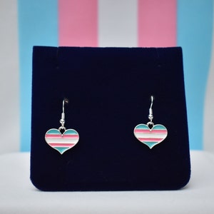 May include: A pair of silver dangle earrings with a heart-shaped charm. The charm is painted with the transgender pride flag colors: light blue, pink, and white.