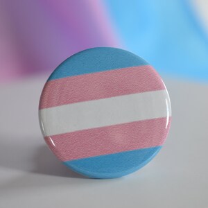 May include: A round button with the transgender pride flag colors: light blue, white, pink, and light blue.