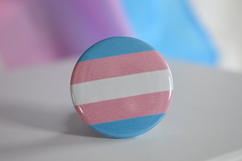 May include: A round button with the transgender pride flag colors: light blue, white, pink, and light blue.