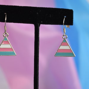 May include: A pair of silver dangle earrings with a triangle-shaped charm. The charm is painted with the transgender pride flag colors: light blue, pink, white, pink, and light blue.