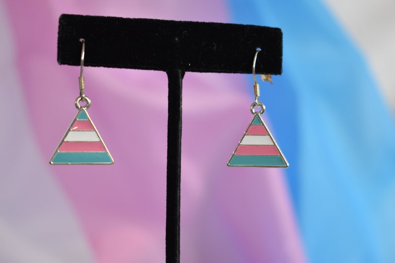 May include: A pair of silver dangle earrings with a triangle-shaped charm. The charm is painted with the transgender pride flag colors: light blue, pink, white, pink, and light blue.