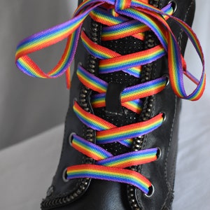 Może przedstawiać: Czarne skórzane buty z tęczowymi sznurowadłami zawiązanymi w krzyżowy wzór.