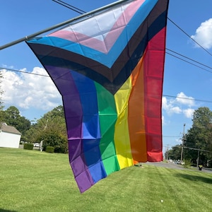 May include: A Progress Pride flag featuring horizontal stripes of red, orange, yellow, green, blue, and violet, with a chevron on the left side containing white, light pink, light blue, brown, and black stripes, mounted on a metal pole.