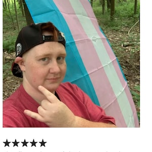 May include: A person is holding a transgender pride flag in a forest. The flag is blue, pink, and white. The person is wearing a red shirt and a black baseball cap.