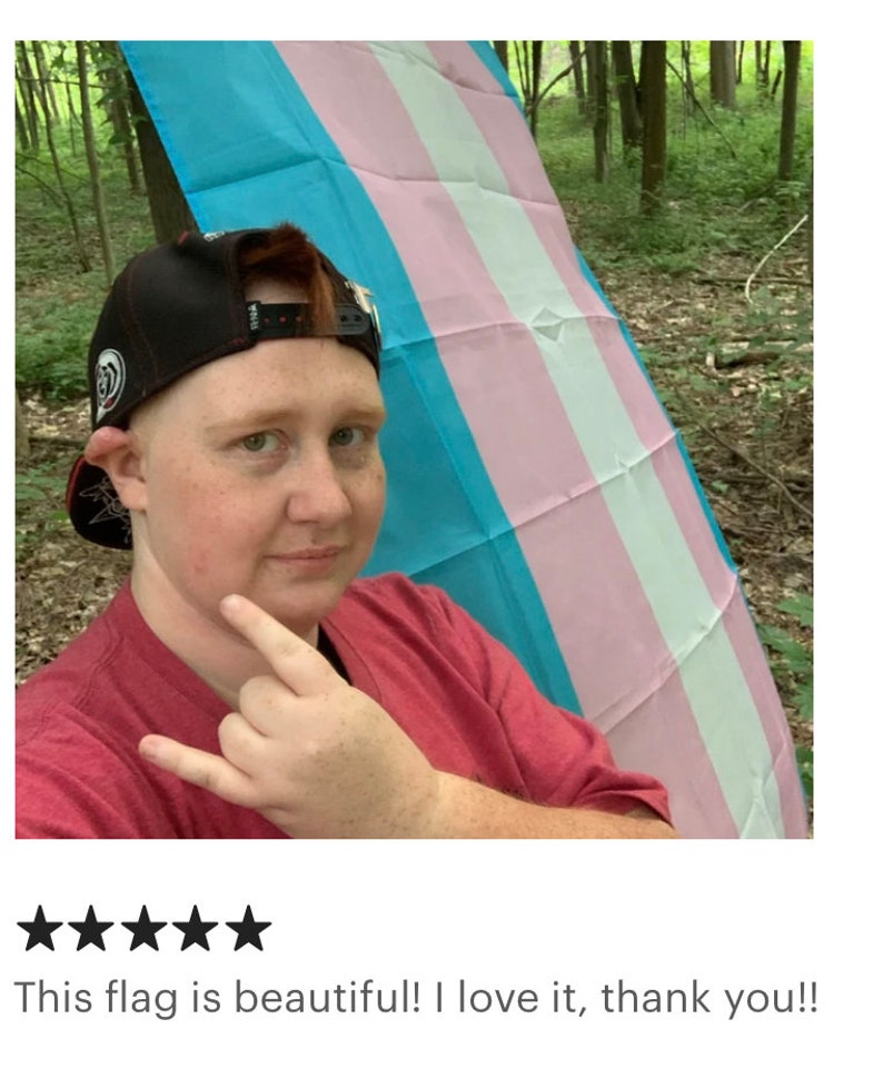 May include: A person is holding a transgender pride flag in a forest. The flag is blue, pink, and white. The person is wearing a red shirt and a black baseball cap.