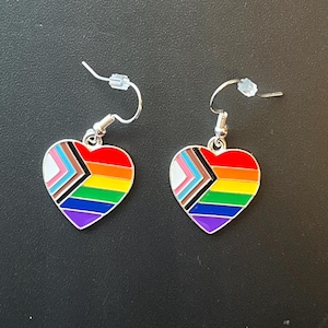 Puede incluir: Un par de pendientes de corazón plateados con rayas arcoíris, negras, marrones, blancas, rosas y azules. Los pendientes cuelgan de ganchos plateados.