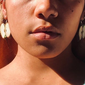 May include: Close-up of a person wearing apple slice earrings. The earrings are made of a light-colored material with a green center, resembling a cross-section of an apple. The earrings hang from silver hooks.