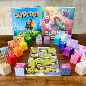 May include: The board game "Cubitos" is displayed with its box, dice, cards, and game board. The box art shows a cartoon character. The dice are in various colours, including red, green, and blue. The game is set on a wooden table.