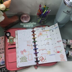 May include: A pink leather planner with a clear plastic cover and a monthly calendar for April. The planner has a rainbow of colorful rings and a variety of stickers and washi tape.
