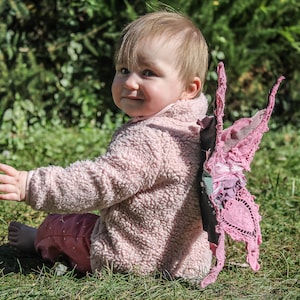 Könnte beinhalten: Ein Baby in einer rosa Fleecejacke und Hose sitzt auf dem Gras und trägt rosa Feenflügel. Die Flügel sind aus Spitze und Stoff gefertigt und haben ein dekoratives Design.