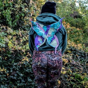 Puede incluir: Una persona con una sudadera con capucha verde oscuro y un gorro negro, con alas de mariposa coloridas en la espalda. Las alas presentan tonos de azul, morado y verde. La persona lleva leggings estampados, de pie en un bosque.