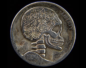 May include: A silver coin with a skull design carved into it. The skull has a floral design on the top of its head.