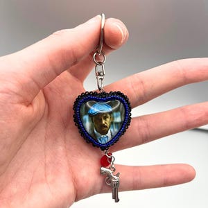May include: Heart-shaped keyring with a portrait of a man in a blue cap. The heart is framed with black and blue beads. A small silver gun charm and a red bead dangle below. The keyring is held by a hand.