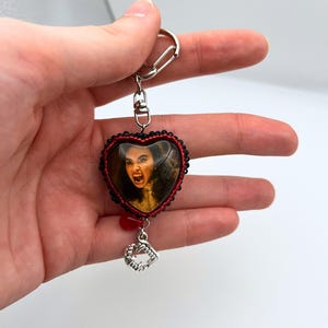 Puede incluir: Llavero en forma de corazón con un retrato de una mujer con cabello oscuro y la boca abierta. El llavero tiene un borde de cuentas negras y rojas, una cuenta roja y un dije plateado de colmillos de vampiro.