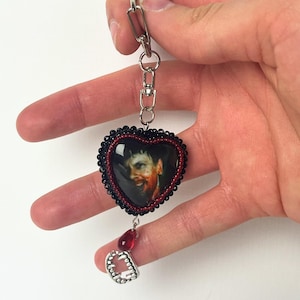 Puede incluir: Un llavero en forma de corazón con un retrato de un hombre con sangre en la cara. El corazón está enmarcado con cuentas negras y rojas. Una cuenta roja en forma de lágrima y un dije de colmillo plateado cuelgan debajo. El llavero se sujeta con una cadena plateada.
