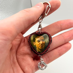 Pode incluir: Chaveiro em forma de coração com uma foto dentro, emoldurado com contas vermelhas e pretas. Um pingente de prata de presas de vampiro pende abaixo. O chaveiro é preso a um fecho de prata.
