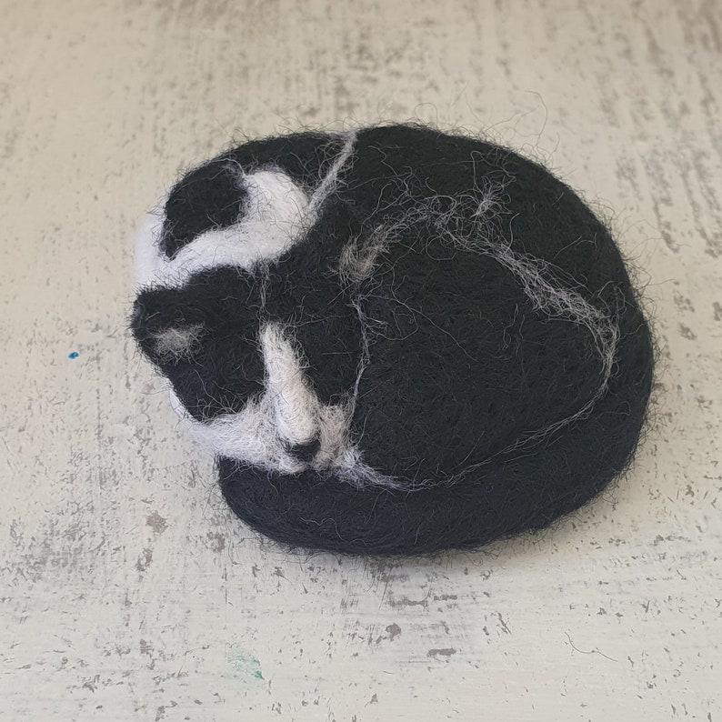 May include: A handmade felted cat figurine in a curled-up sleeping position. The cat is primarily black with white markings on its face and body. The figurine is round and rests on a light-colored surface.