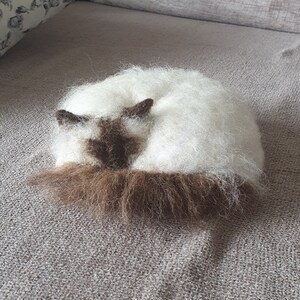 May include: A handmade, felted cat figurine in a curled-up position. The cat has a white body with brown markings on its face, ears, and tail. The fur appears soft and fluffy, giving the figurine a realistic appearance. The cat is resting on a gray surface.