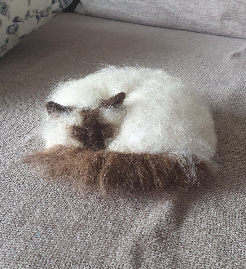 May include: A handmade, felted cat figurine in a curled-up position. The cat has a white body with brown markings on its face, ears, and tail. The fur appears soft and fluffy, giving the figurine a realistic appearance. The cat is resting on a gray surface.