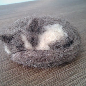 May include: A handmade felted cat figurine in a curled-up sleeping position. The cat is primarily gray with a white patch on its chest and belly. The figurine is resting on a wooden surface.