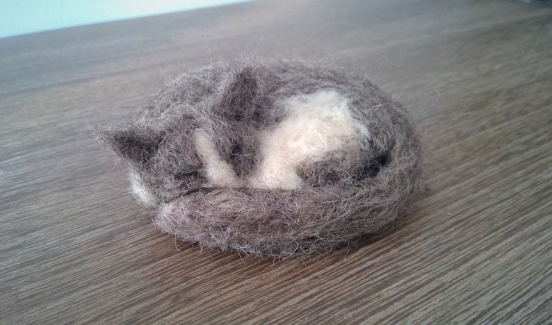 May include: A handmade felted cat figurine in a curled-up sleeping position. The cat is primarily gray with a white patch on its chest and belly. The figurine is resting on a wooden surface.