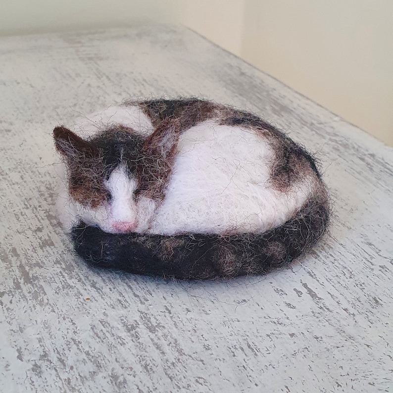 May include: A handmade felted cat figurine curled up in a sleeping position. The cat has white fur with brown and black markings, a pink nose, and a black tail. The figurine is on a light gray surface.