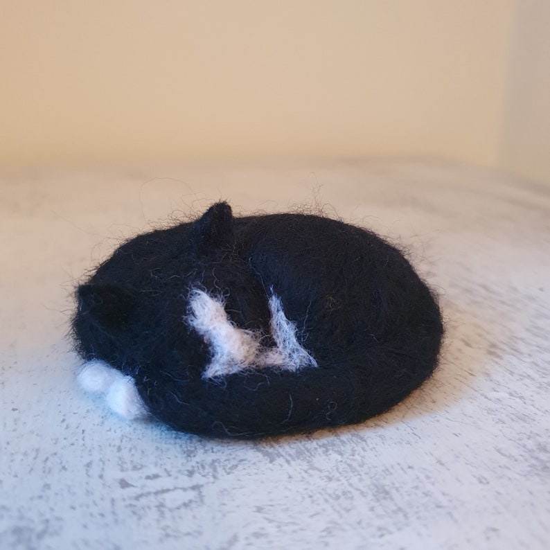 May include: A handmade felted cat figurine in a curled-up sleeping position. The cat is primarily black with white markings on its chest and paws. The figurine is resting on a light-colored surface.