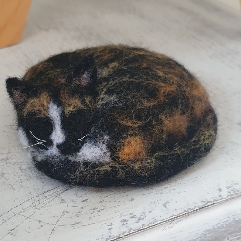May include: A felted cat figurine in a curled sleeping position. The cat has a black, brown, and orange coat with white markings on its face. The cat's eyes are closed, and it has white whiskers.