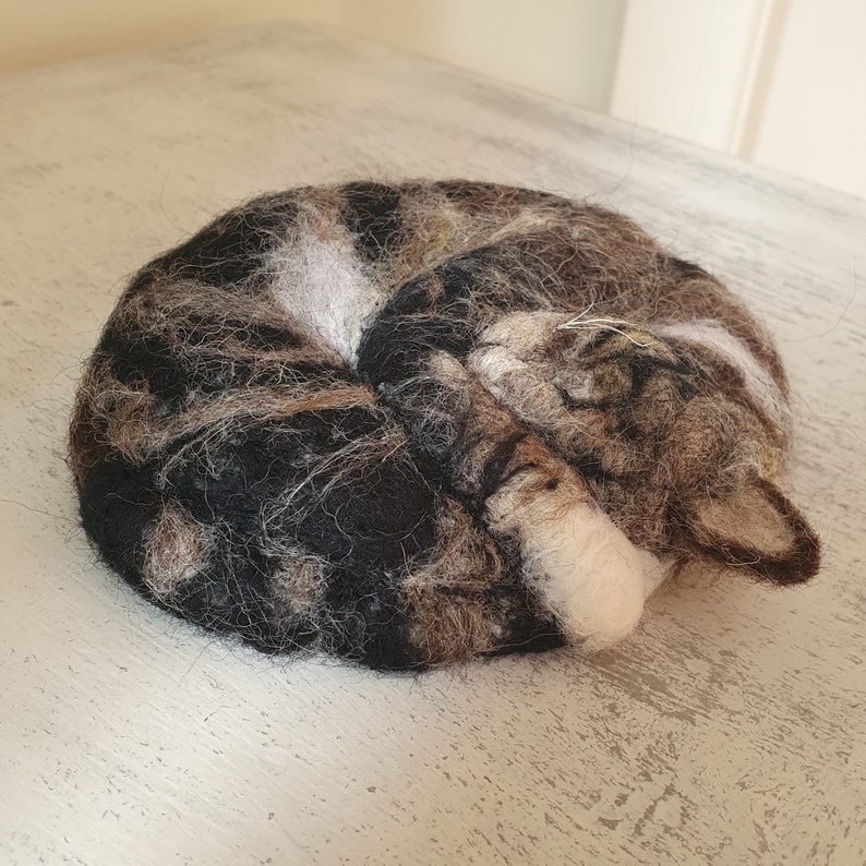 May include: A felted cat figurine curled up in a ball. The cat's fur is a mix of brown, black, and white, with a white patch on its belly and a white paw. The cat is in a sleeping position.