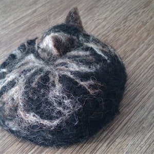 May include: A handmade felted cat figurine in a curled-up position. The cat is primarily black with white and gray markings, and a small, upright ear is visible. The texture of the felt is visible, and the figurine rests on a wooden surface.