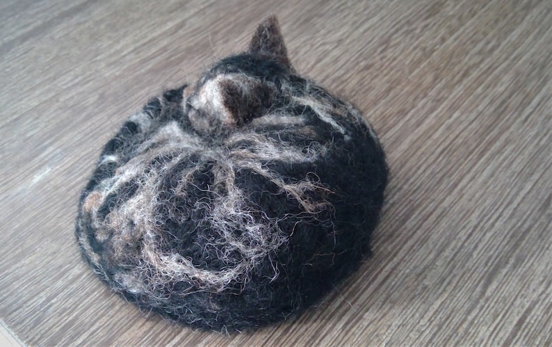 May include: A handmade felted cat figurine in a curled-up position. The cat is primarily black with white and gray markings, and a small, upright ear is visible. The texture of the felt is visible, and the figurine rests on a wooden surface.