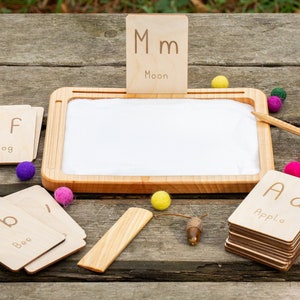 Pode incluir: Conjunto educativo de madeira com cart&otilde;es do alfabeto, uma bandeja com inser&ccedil;&atilde;o branca e bolas de feltro coloridas. Os cart&otilde;es exibem letras e palavras como "Ma&ccedil;&atilde;", "Abelha", "Sapo" e "Lua". Uma caneta de madeira e bolotas tamb&eacute;m est&atilde;o inclu&iacute;das.
