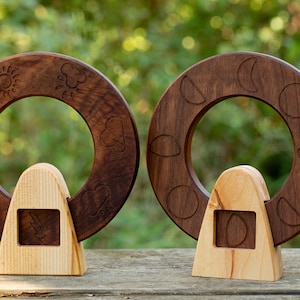May include: Two wooden rings with carved designs. The ring on the left has sun, moon, and weather symbols. The ring on the right has moon phases. Both rings are mounted on wooden stands.