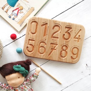 Op de afbeelding: Houten cijfertraceerbord met gesneden cijfers 0-9. Het bord is gemaakt van lichtgekleurd hout en bevat pijlen voor elke cijfer. Een pop, een mand met kleurrijke ballen en een kinderboek staan ook in de afbeelding.