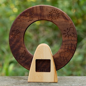 May include: A wooden weather wheel with carved weather symbols, including a sun, cloud, snowflake, lightning bolt, wind, and heart. The wheel is dark brown and sits on a light-colored wooden base with a cloud and rain symbol.