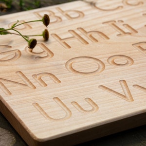 Op de afbeelding: Houten alfabet traceerbord met hoofdletters en kleine letters in het oppervlak gegraveerd. Het bord is gemaakt van lichtgekleurd hout en heeft een natuurlijke afwerking. Een takje groen met kleine bruine knoppen ligt op het bord.