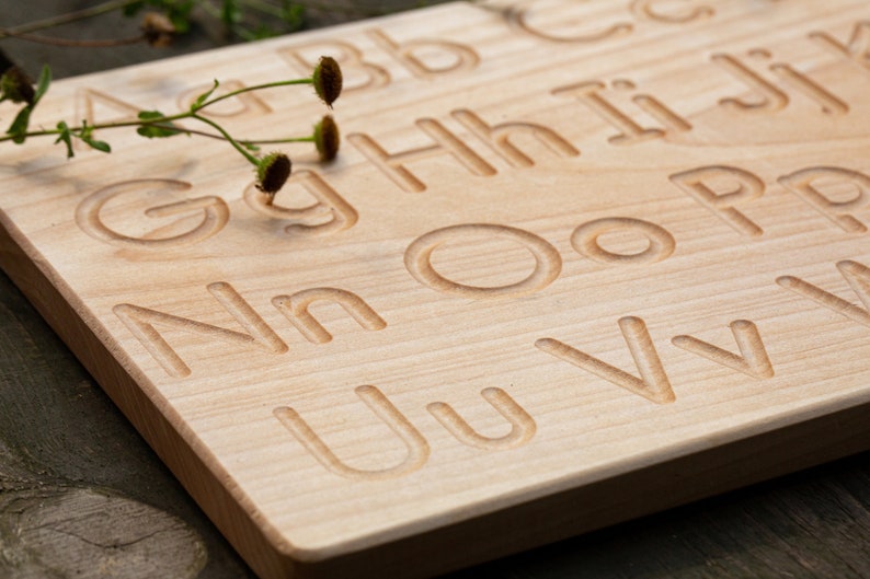 Op de afbeelding: Houten alfabet traceerbord met hoofdletters en kleine letters in het oppervlak gegraveerd. Het bord is gemaakt van lichtgekleurd hout en heeft een natuurlijke afwerking. Een takje groen met kleine bruine knoppen ligt op het bord.