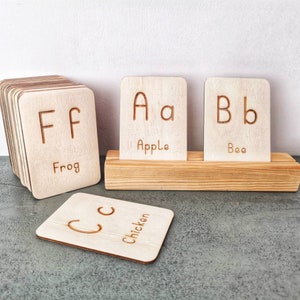 Pode incluir: Cart&otilde;es de alfabeto em madeira com letras gravadas e palavras correspondentes. Os cart&otilde;es apresentam as letras F, A, B e C, com as palavras "Frog", "Apple", "Bee" e "Chicken". Os cart&otilde;es s&atilde;o exibidos em um suporte de madeira.