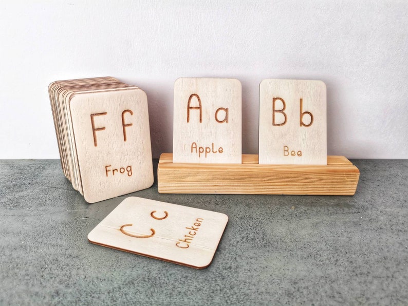 Pode incluir: Cart&otilde;es de alfabeto em madeira com letras gravadas e palavras correspondentes. Os cart&otilde;es apresentam as letras F, A, B e C, com as palavras "Frog", "Apple", "Bee" e "Chicken". Os cart&otilde;es s&atilde;o exibidos em um suporte de madeira.