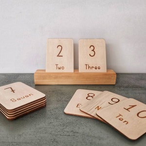 May include: Wooden number cards with engraved numerals and words. The cards are displayed on a wooden stand and stacked. Numbers 2, 3, 7, 8, 9, and 10 are visible. Educational toy for learning numbers.