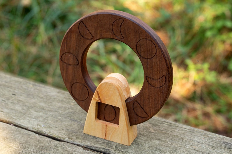May include: A wooden moon phase ornament with a dark brown ring and a light brown base. The ring has eight different moon phases carved into it.