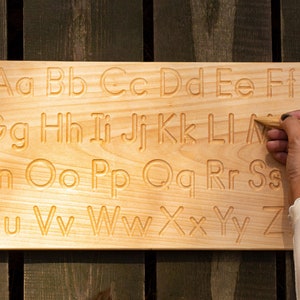 Op de afbeelding: Houten alfabetbord met hoofdletters en kleine letters. Een hand gebruikt een houten stylus om de letter "L" te traceren. Het bord is gemaakt van lichtgekleurd hout en staat tegen een donkere houten achtergrond.