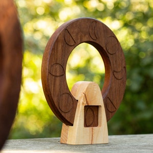May include: A wooden sculpture of a Ferris wheel with a brown wheel and a light brown base. The wheel has a series of crescent moon shapes carved into it.