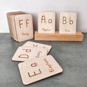 Pode incluir: Cart&otilde;es de alfabeto de madeira com letras gravadas e palavras correspondentes. Os cart&otilde;es s&atilde;o exibidos em um suporte de madeira e empilhados. As letras incluem A, B, D, E e F, com palavras como Apple, Bee, Daisy, Elephant e Frog.