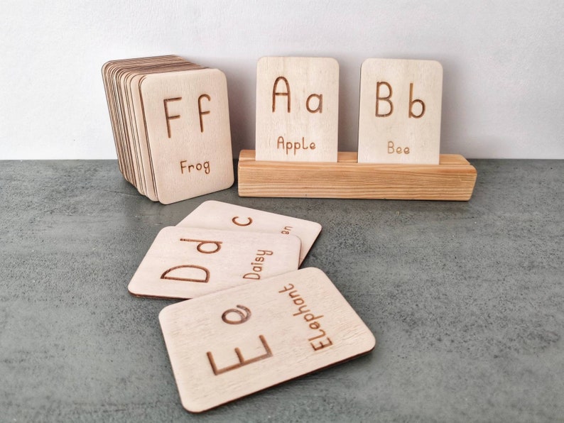 Pode incluir: Cart&otilde;es de alfabeto de madeira com letras gravadas e palavras correspondentes. Os cart&otilde;es s&atilde;o exibidos em um suporte de madeira e empilhados. As letras incluem A, B, D, E e F, com palavras como Apple, Bee, Daisy, Elephant e Frog.