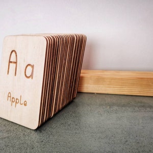 Pode incluir: Cart&otilde;es de mem&oacute;ria de madeira com a letra "A" e a palavra "Apple" gravadas na frente. Os cart&otilde;es est&atilde;o empilhados e encostados em um bloco de madeira. O fundo &eacute; uma superf&iacute;cie cinza claro.