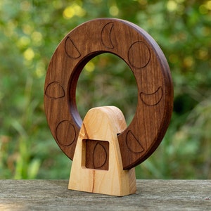 May include: A wooden moon phase wheel with a carved wooden base. The wheel is made of dark wood and has eight different moon phases carved into it. The base is made of light wood and has a small opening in the front.