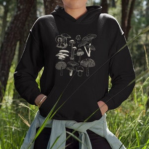 May include: Black hooded sweatshirt with a cream-coloured illustration of different mushrooms. The design showcases various fungi, with diverse shapes and sizes. The hoodie has a drawstring hood and a front pocket.